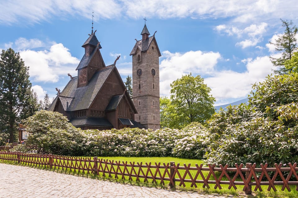 A historic building with a tall tower and steeply pitched roofs, surrounded by lush greenery and a wooden fence.