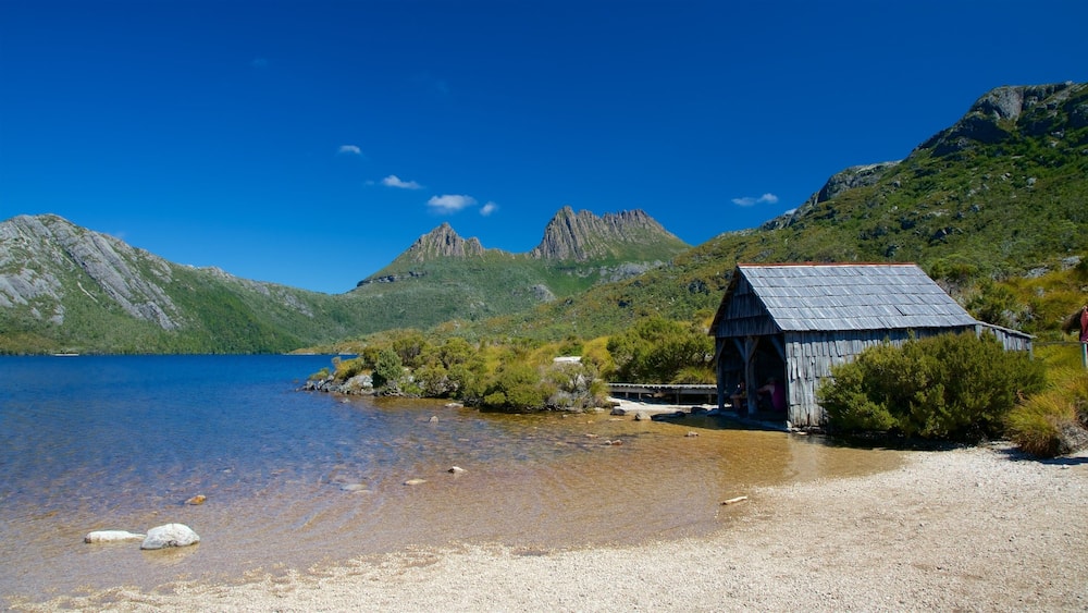 A small wooden structure by a clear lake with mountains in the background.