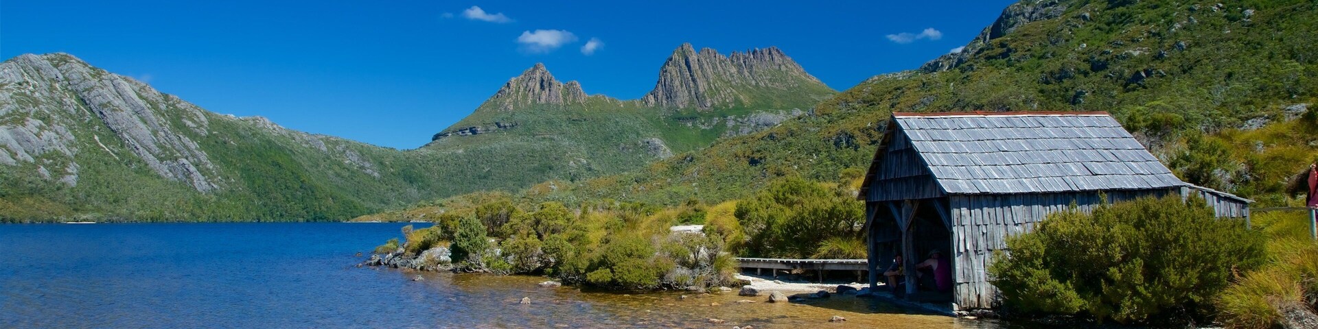 Una pequeña estructura de madera junto a un lago cristalino con montañas al fondo.