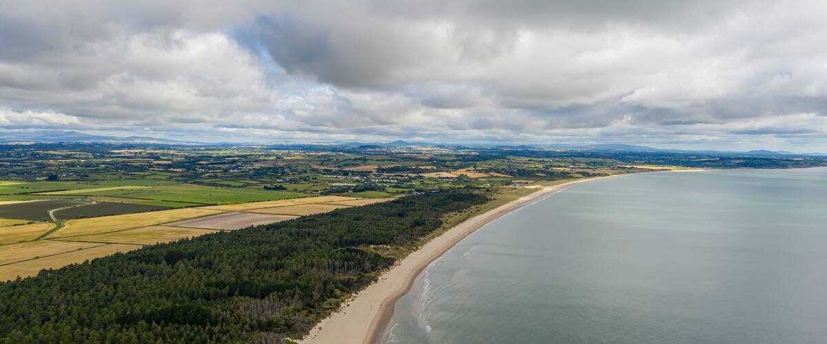 An aerial view of a coastal landscape with a sandy beach, dense forest, and a large body of water under a cloudy sky.
