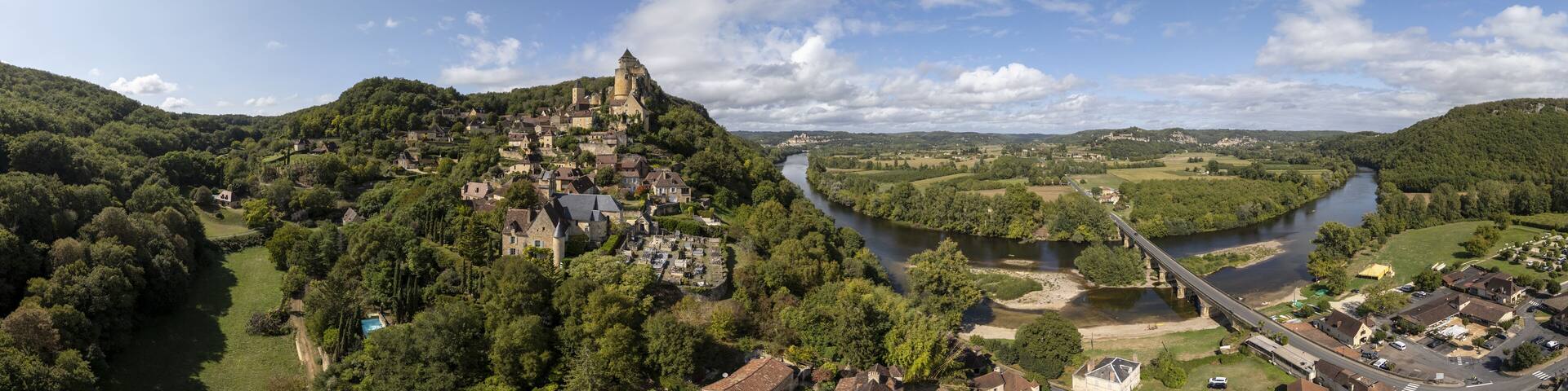 Aerial view of a dramatic castle perched atop a forested hill, overlooking a winding river under a partly cloudy sky, Beynac-et-Cazenac, Dordogne, France.