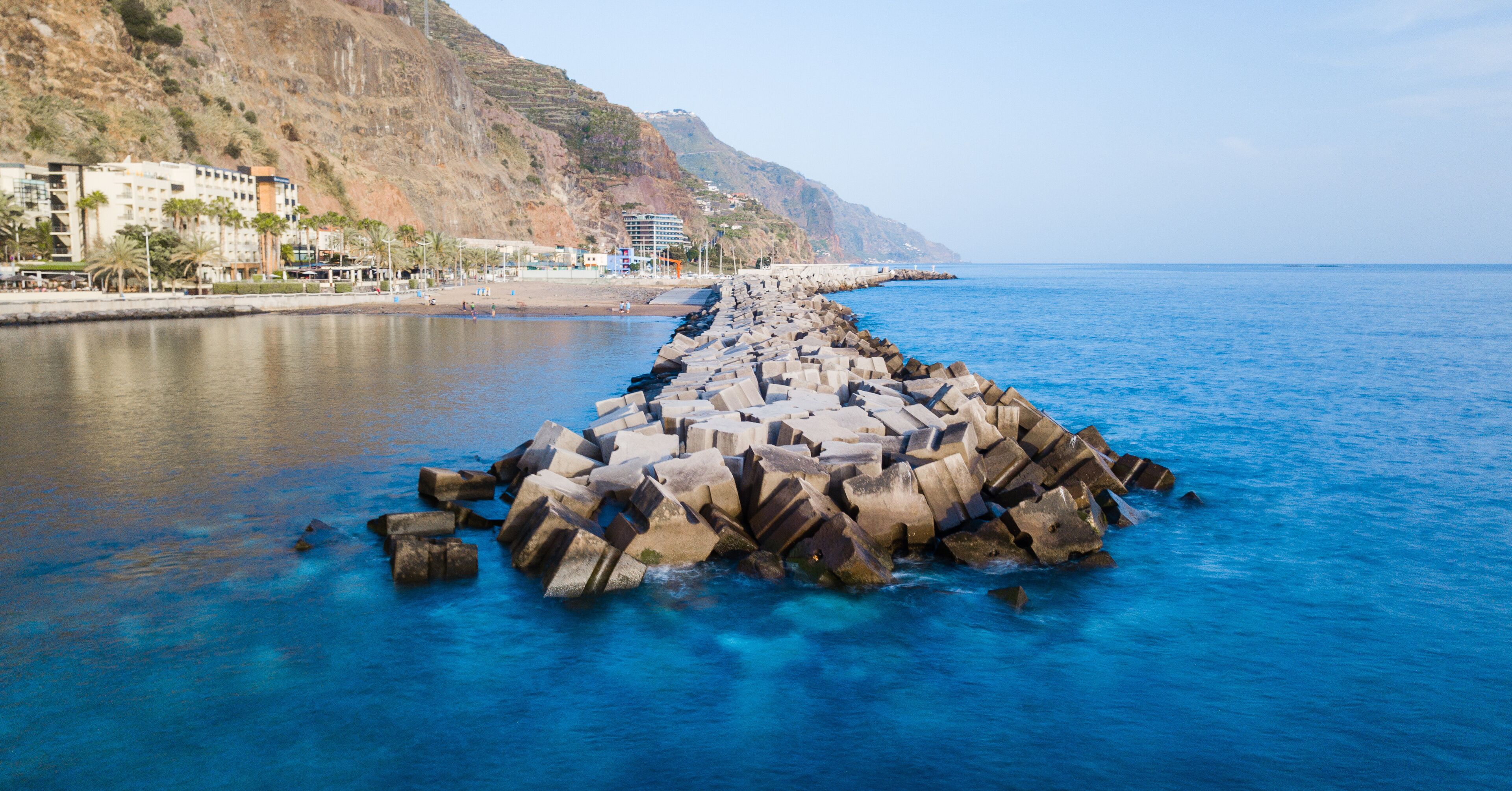 A rocky breakwater extending into the sea with buildings on the shore.