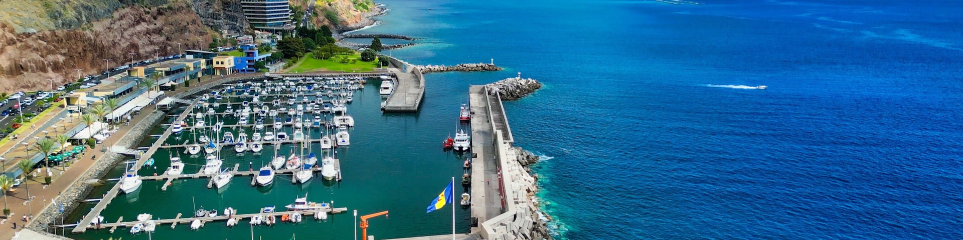 Una marina costera con barcos amarrados, una playa de arena y montañas al fondo.