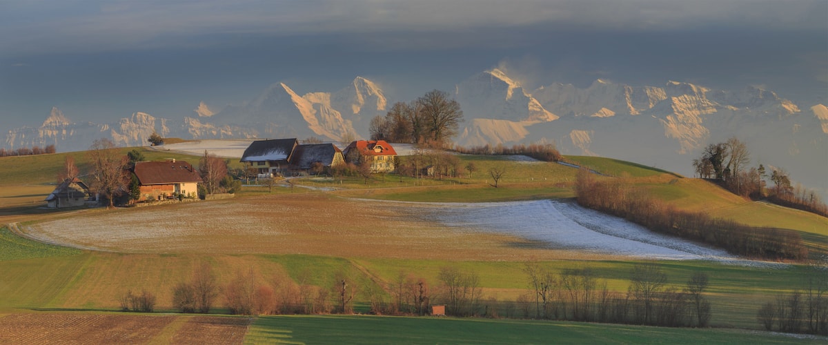 A rural landscape with rolling hills, a farmhouse, and snow-capped mountains in the background.