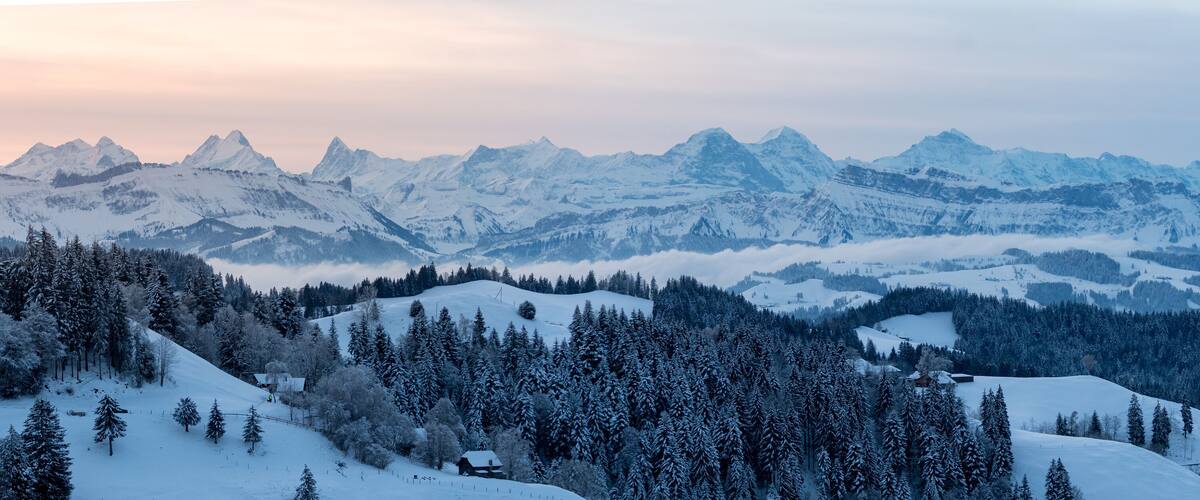 A snow-covered mountain range with dense forests and a few small buildings.