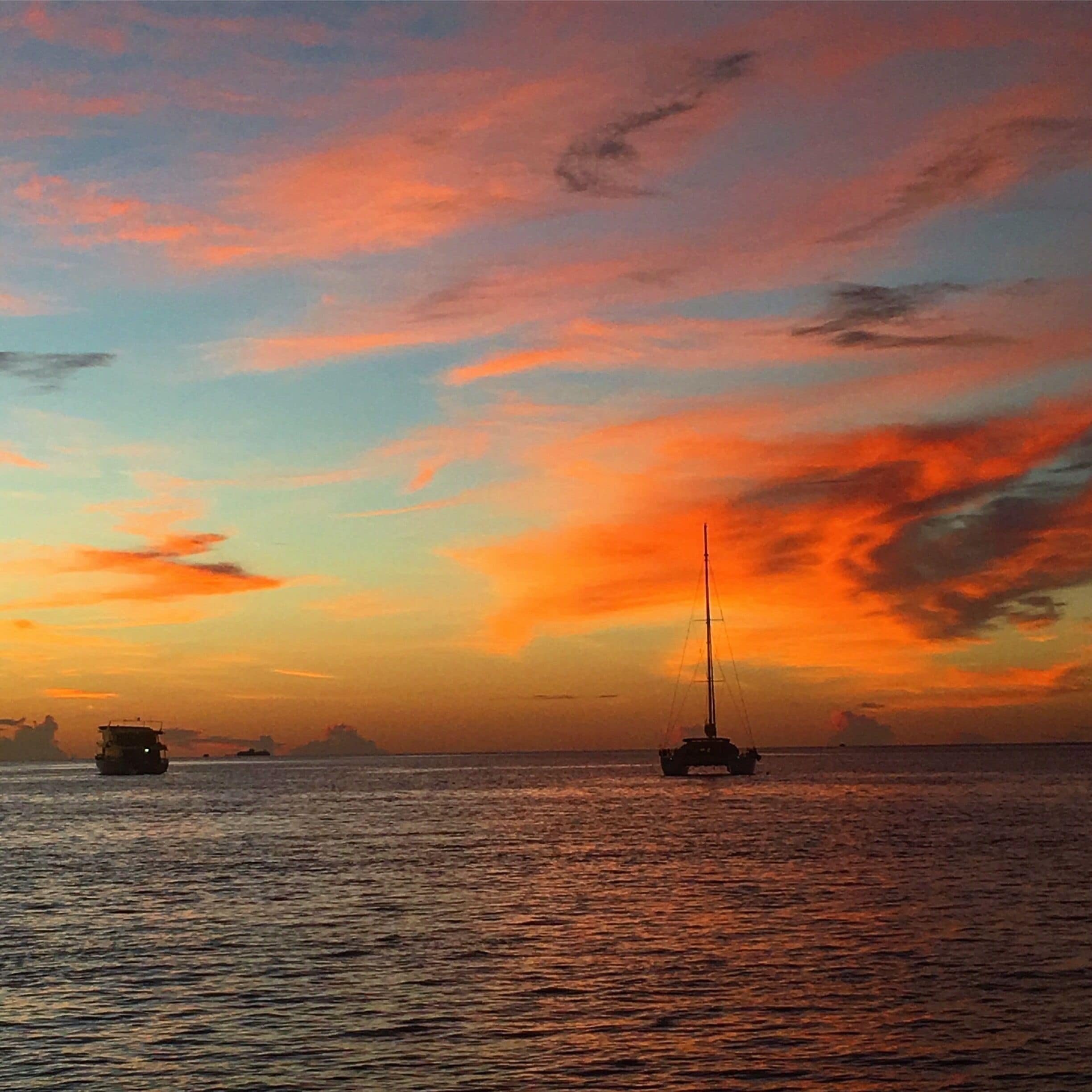 A sunset over a calm sea with a sailboat and a ship in the distance.