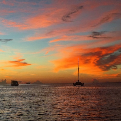 Un coucher de soleil sur une mer calme, avec un voilier et un navire à l’horizon.