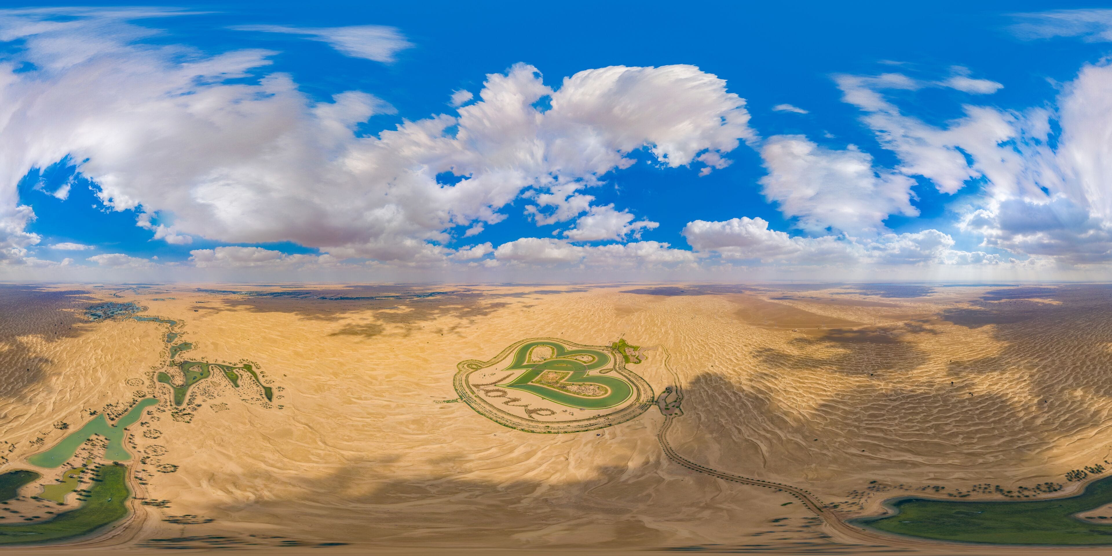 A desert landscape with a winding road and green areas, under a partly cloudy sky.