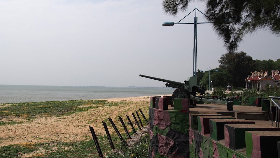 A coastal area with a military cannon, a beach, and a distant bridge.