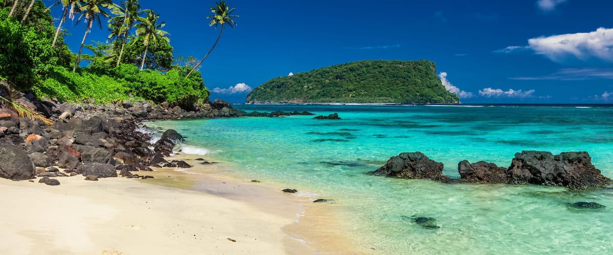A tropical beach with clear turquoise waters, sandy shore, and lush greenery including palm trees.