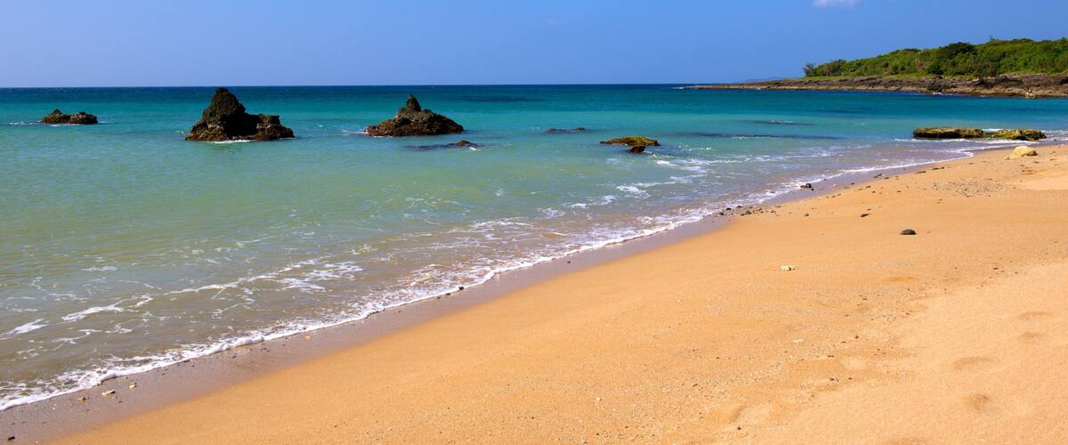 A sandy beach with clear turquoise water, rocky outcrops, and a distant green hill.