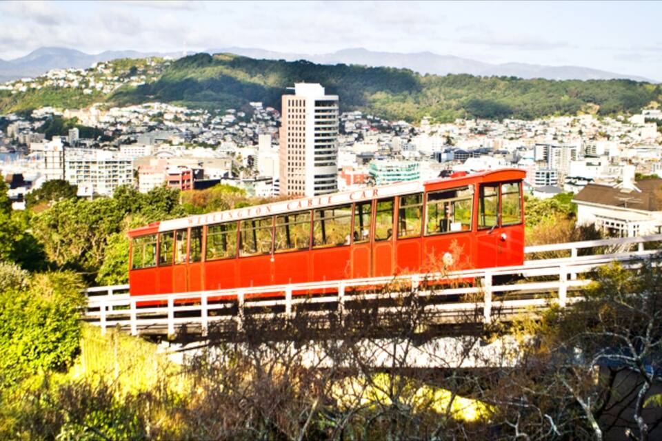 A red cable car on a bridge with a cityscape and mountains in the background.