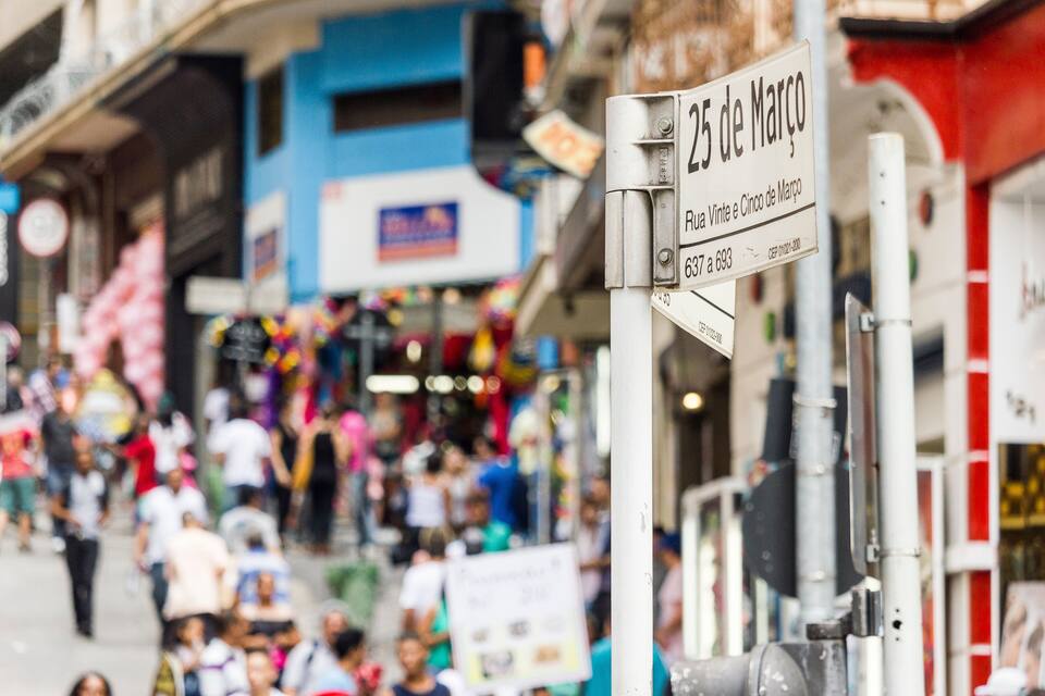 A busy street with a sign indicating '25 de Março' and 'Rua Vinte e Cinco de Março'.
