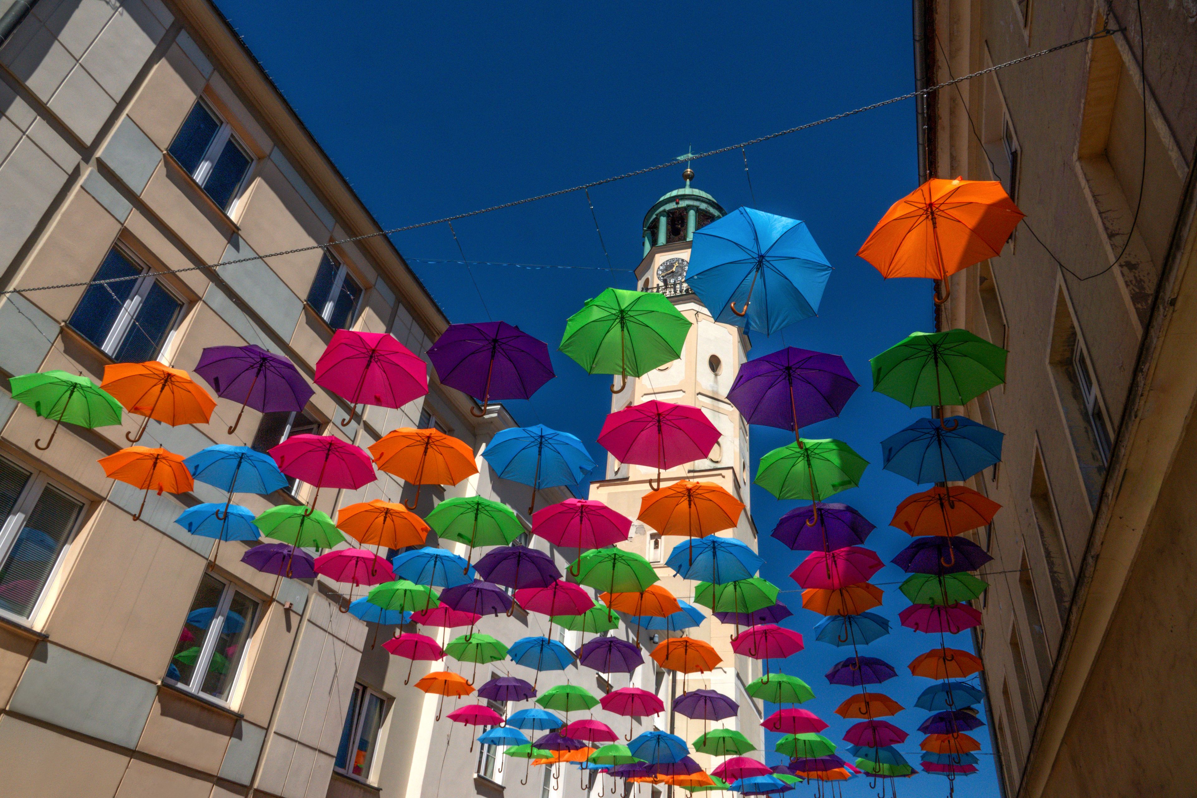 Multi-colored umbrellas hanging between buildings leading towards an old white city clock tower. Olesnica, Poland