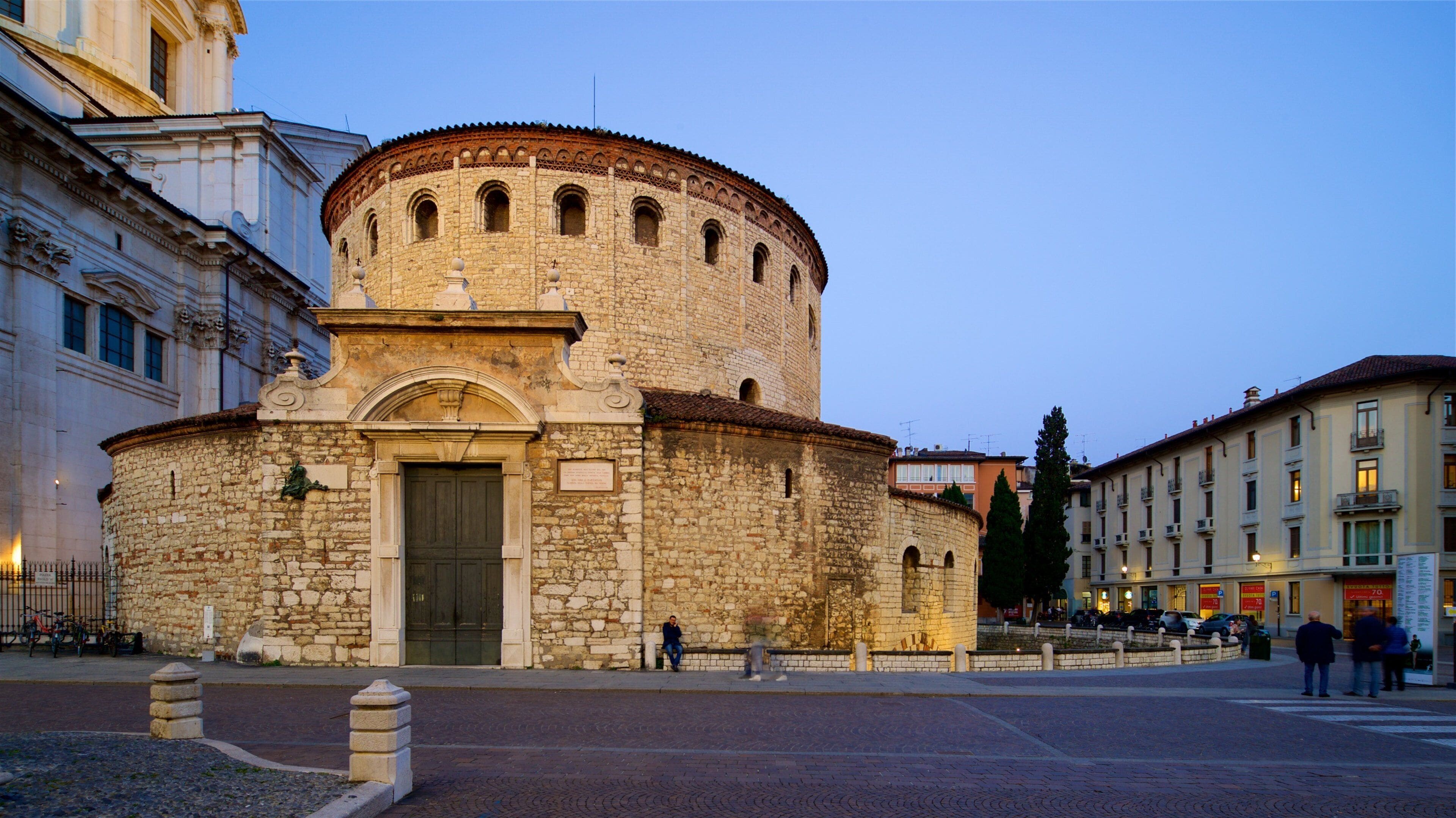 A historic stone building with a round tower and arched entrance, surrounded by modern buildings and a paved square.