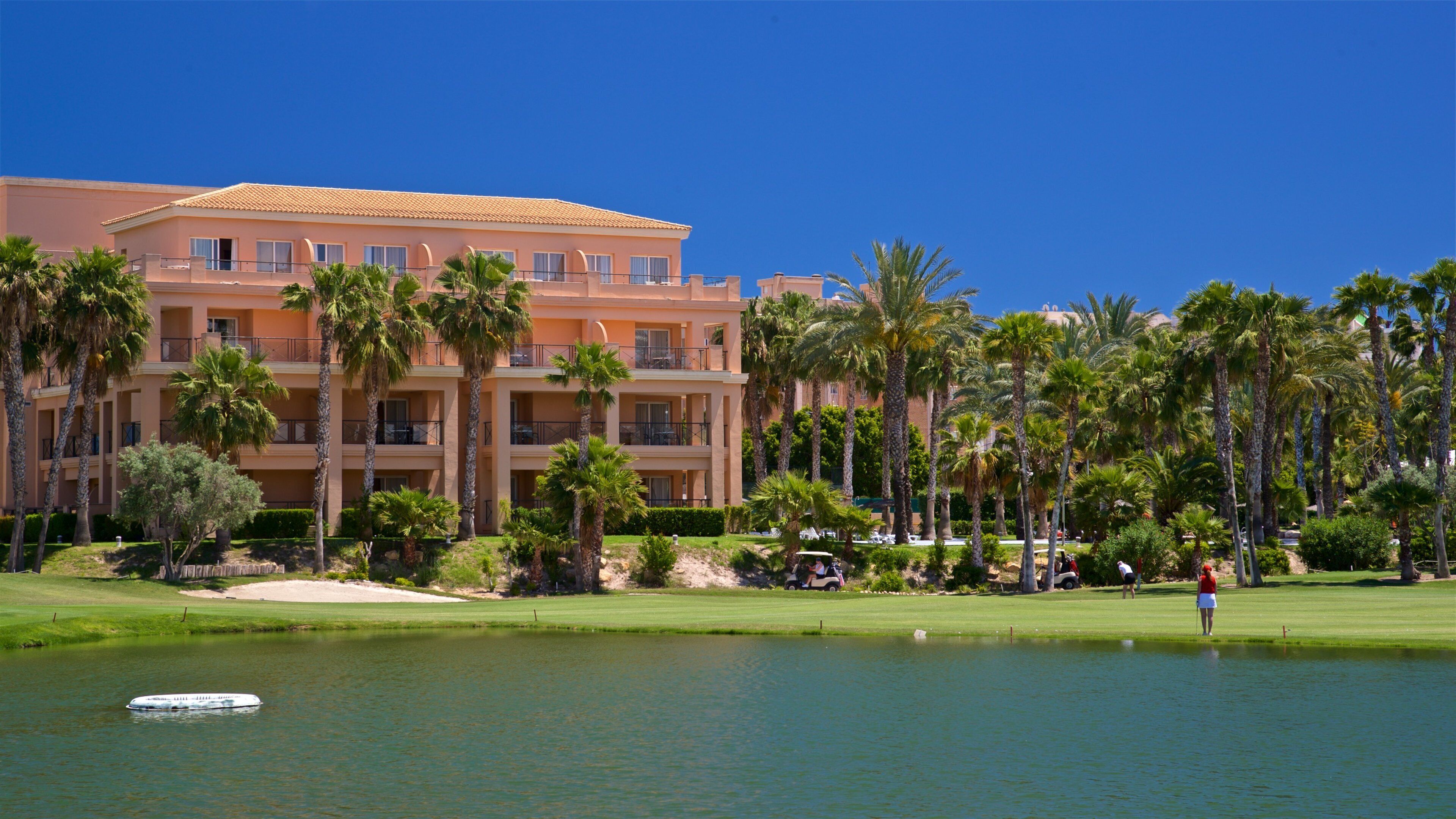 A golf course with a water hazard, palm trees, and a hotel building.
