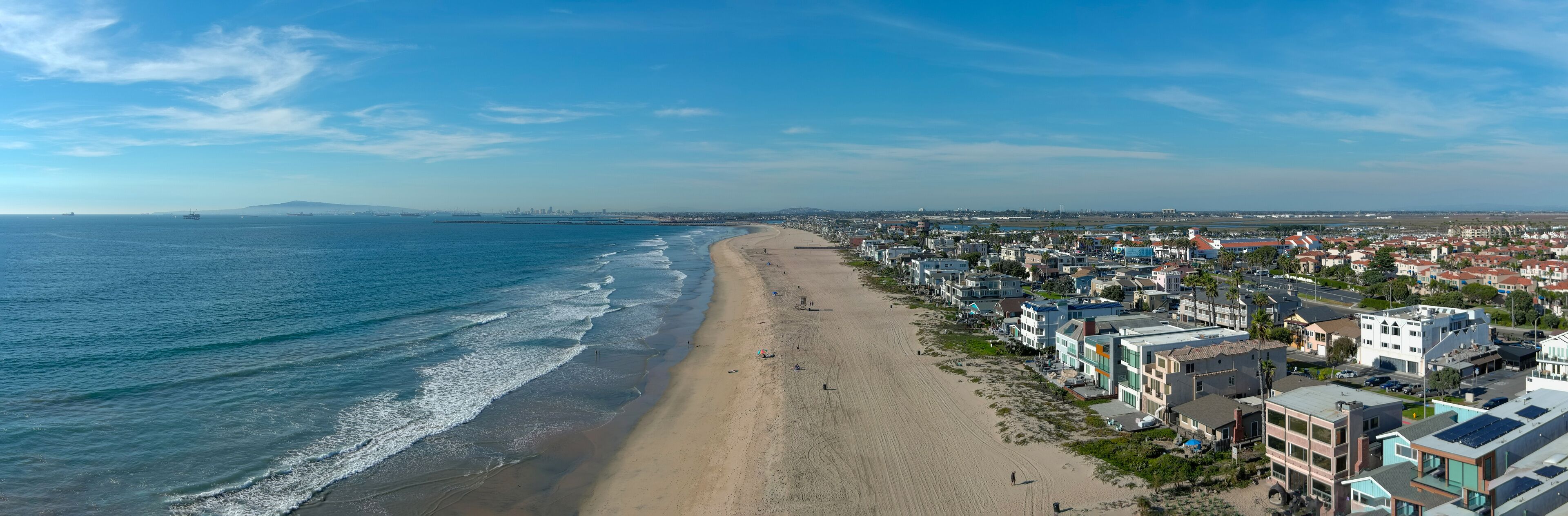 aerial shot of the coastline with blue ocean water and homes along the sand on the beach, cars driving on the street and blue sky with clouds in Huntington Beach California USA