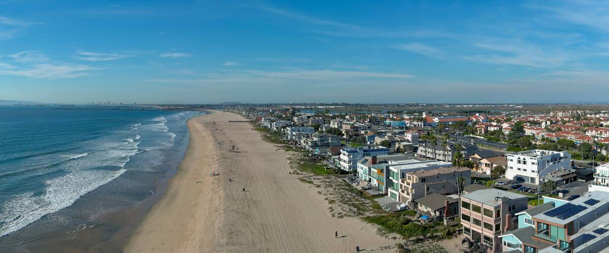aerial shot of the coastline with blue ocean water and homes along the sand on the beach, cars driving on the street and blue sky with clouds in Huntington Beach California USA
