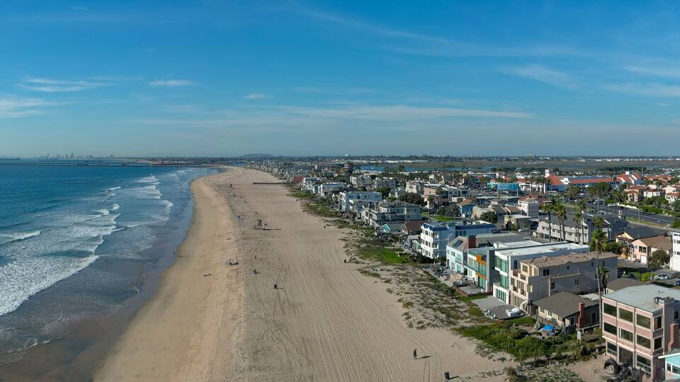 aerial shot of the coastline with blue ocean water and homes along the sand on the beach, cars driving on the street and blue sky with clouds in Huntington Beach California USA