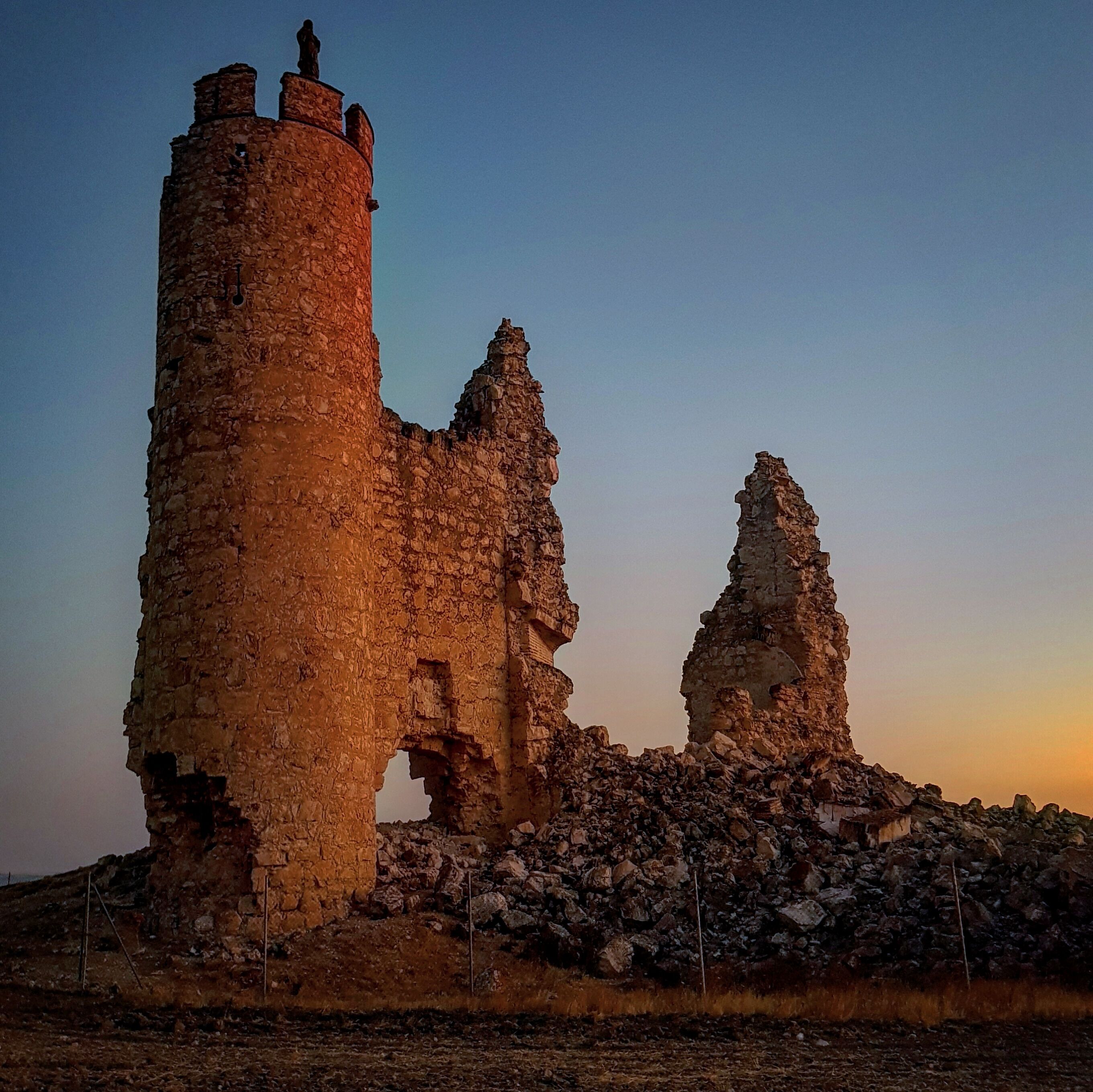 A ruined stone tower with a statue on top, surrounded by rubble and a clear sky.