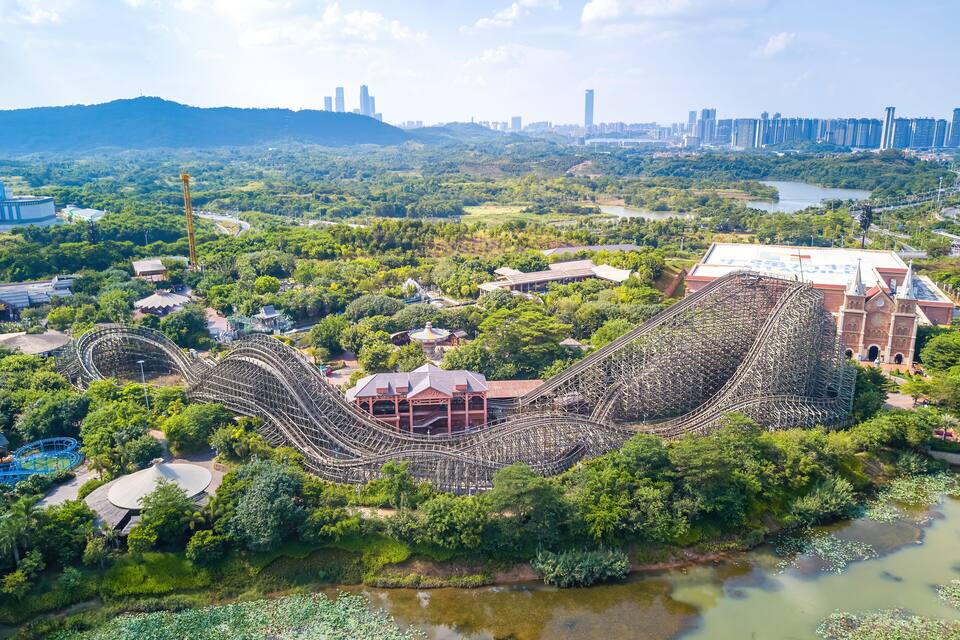 Una montaña rusa en un parque verde exuberante, con un horizonte urbano de fondo.