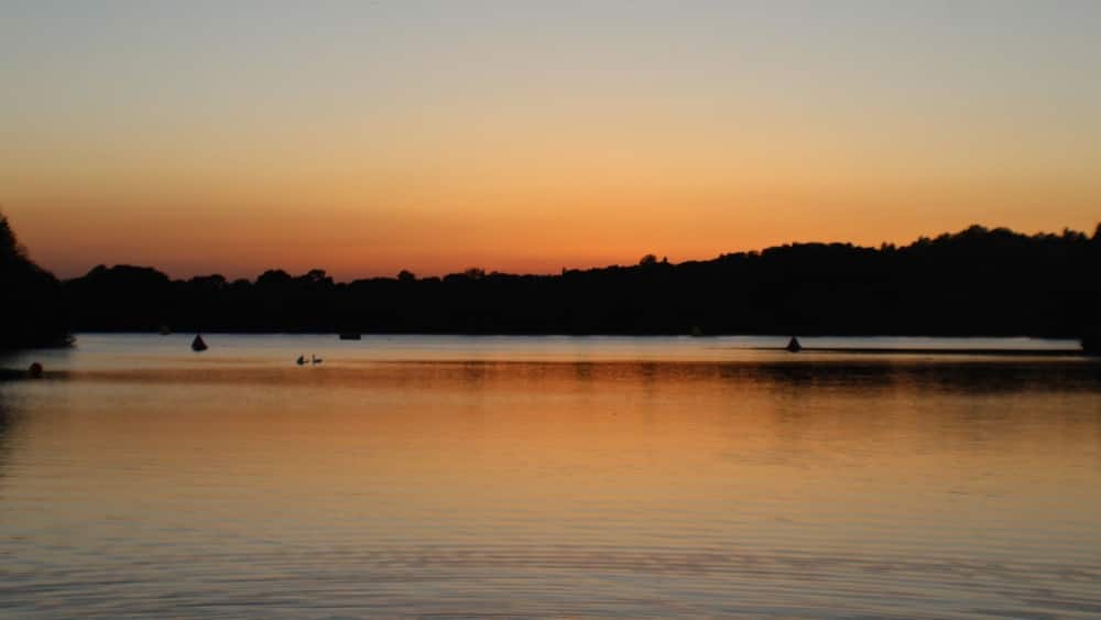 A calm lake at sunset with a silhouette of trees on the horizon and a few boats on the water.
