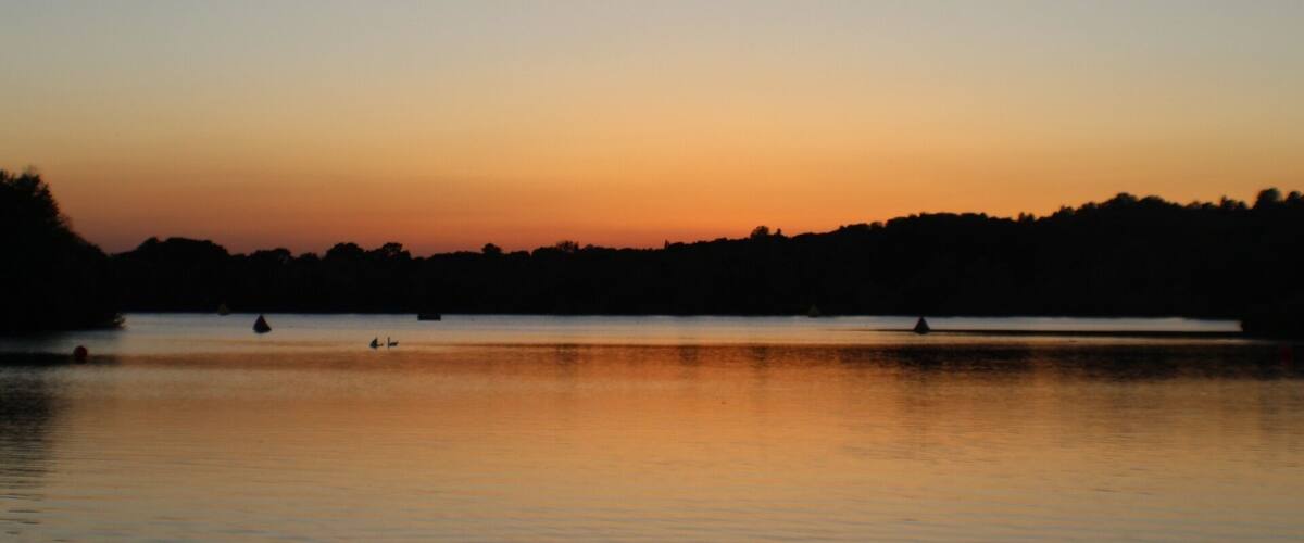 A calm lake at sunset with a silhouette of trees on the horizon and a few boats on the water.