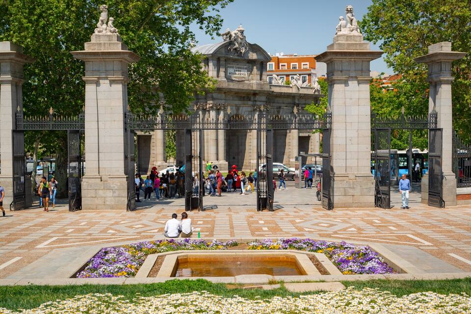 A historic gate with statues, surrounded by a plaza with patterned paving and flower beds.
