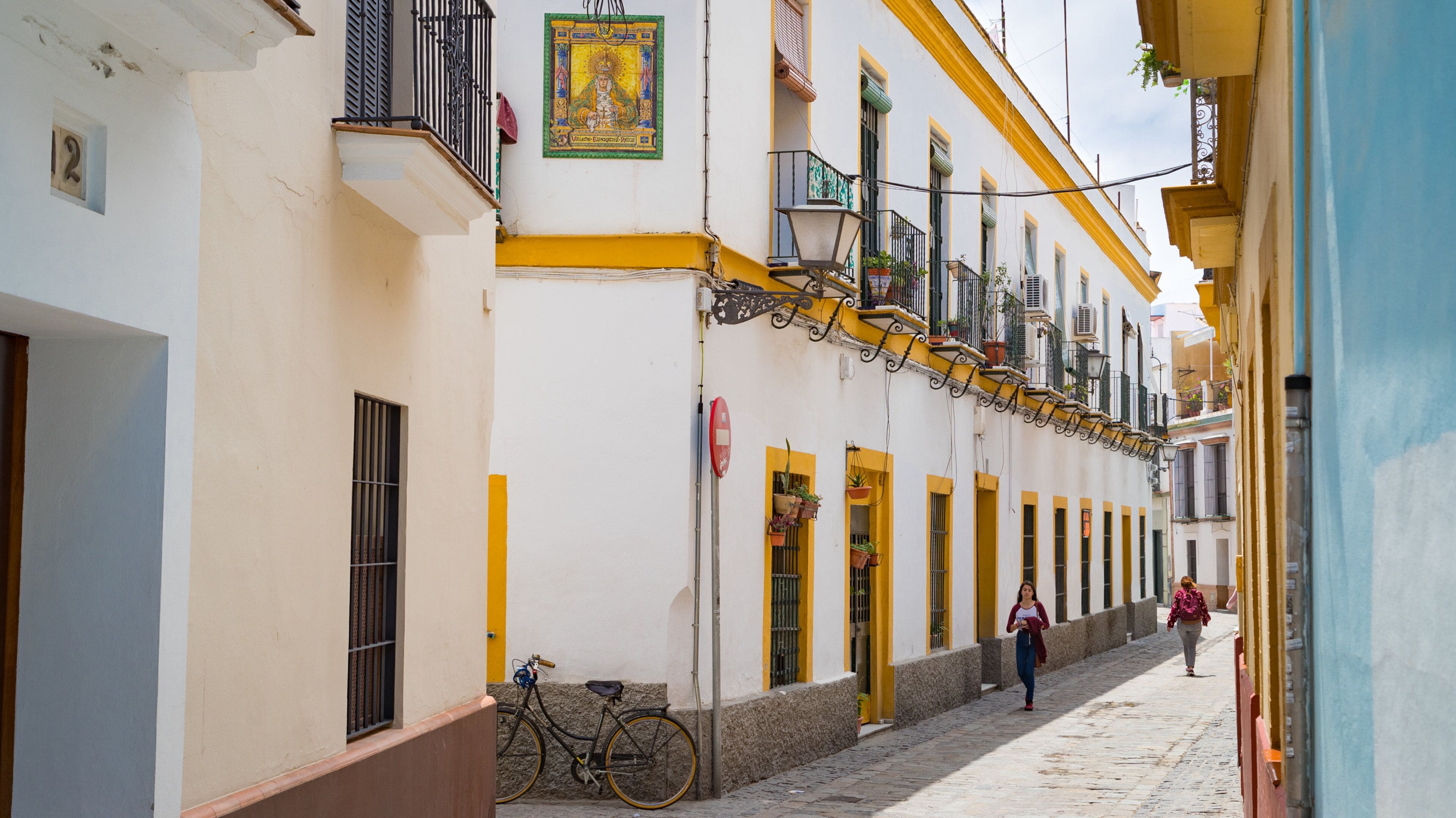 Uma rua de paralelepípedos estreita, ladeada por edifícios brancos com detalhes amarelos, uma bicicleta e uma pessoa caminhando.