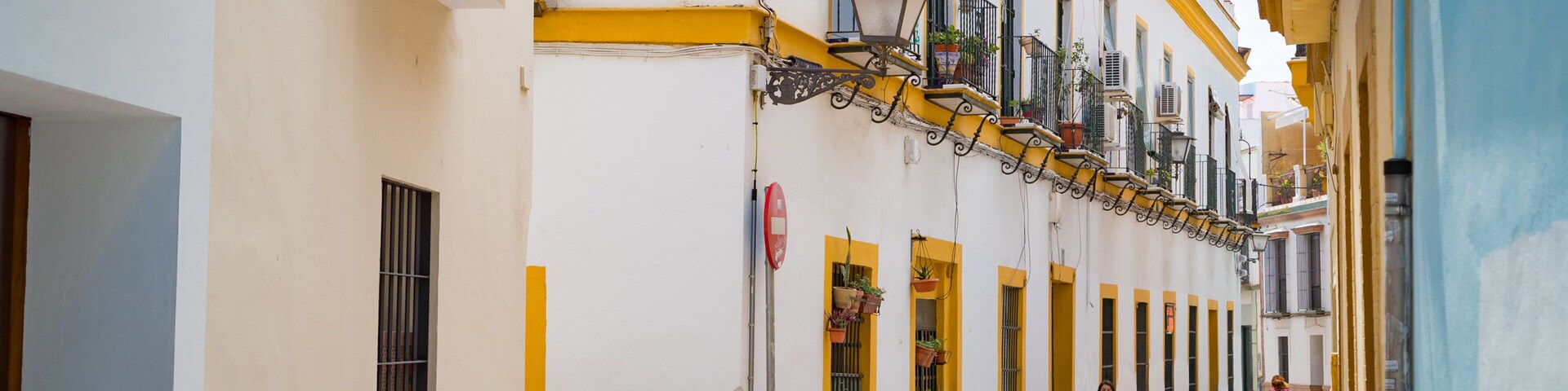Uma rua de paralelepípedos estreita, ladeada por edifícios brancos com detalhes amarelos, uma bicicleta e uma pessoa caminhando.