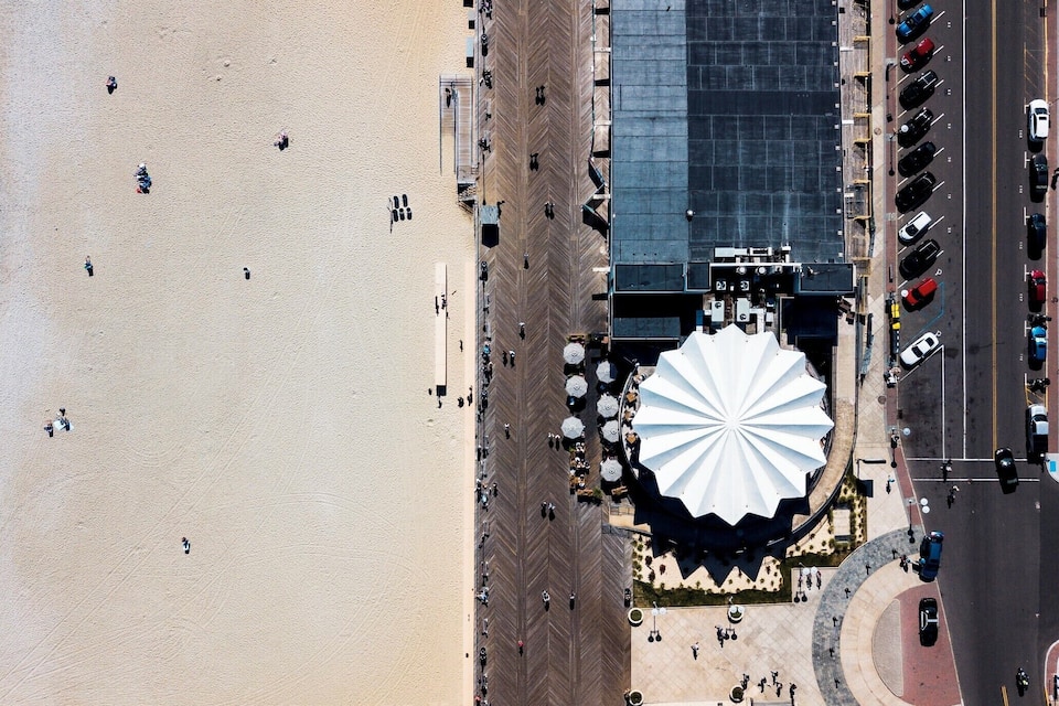 An aerial view of a beach with a large white structure, likely a pavilion, and a parking lot with cars.