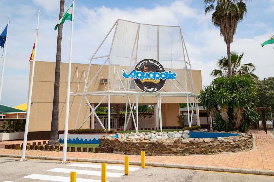 A building with a sign reading "Parque Acuático Aquopolis Sevilla" surrounded by palm trees and flags.