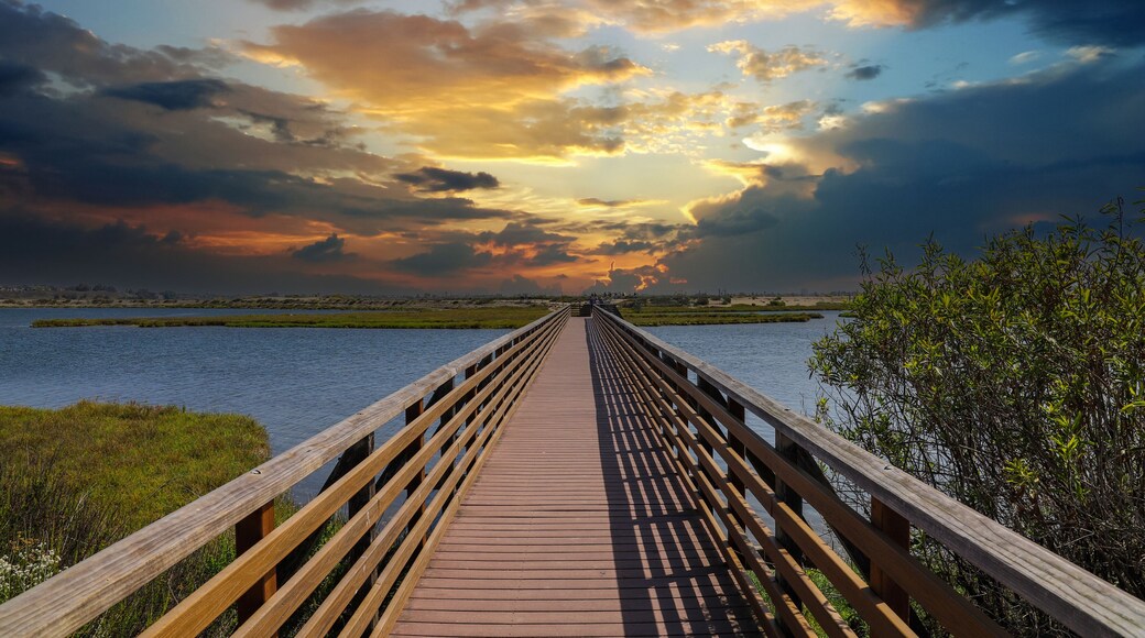 a long brown wooden bridge over a lush green marsh surrounded by vast blue ocean water with powerful clouds at sunset at Bolsa Chica Ecological Reserve in Huntington Beach California USA