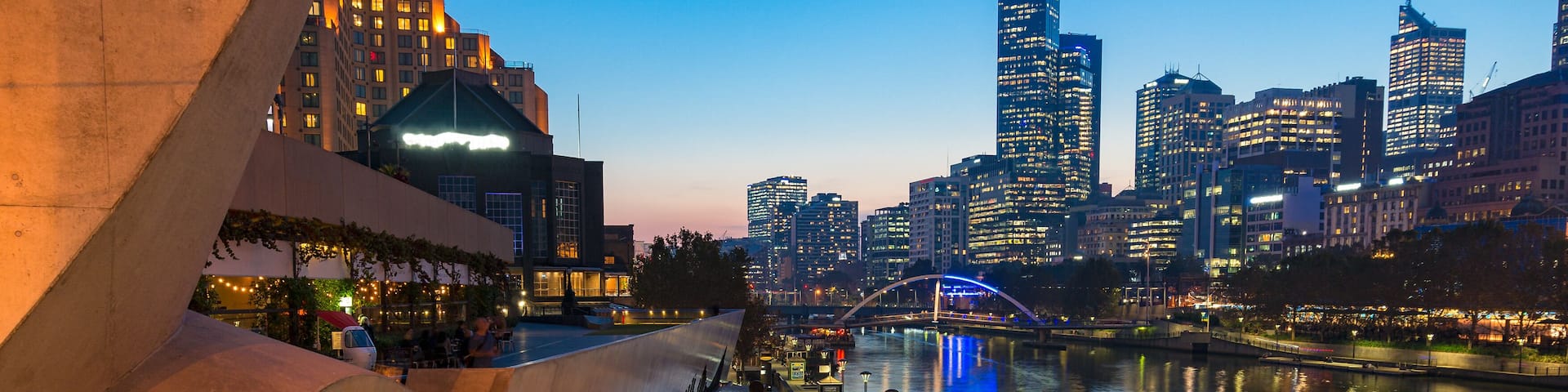 A waterfront promenade with a modern architectural structure, a river reflecting city lights, and a bridge in the background.