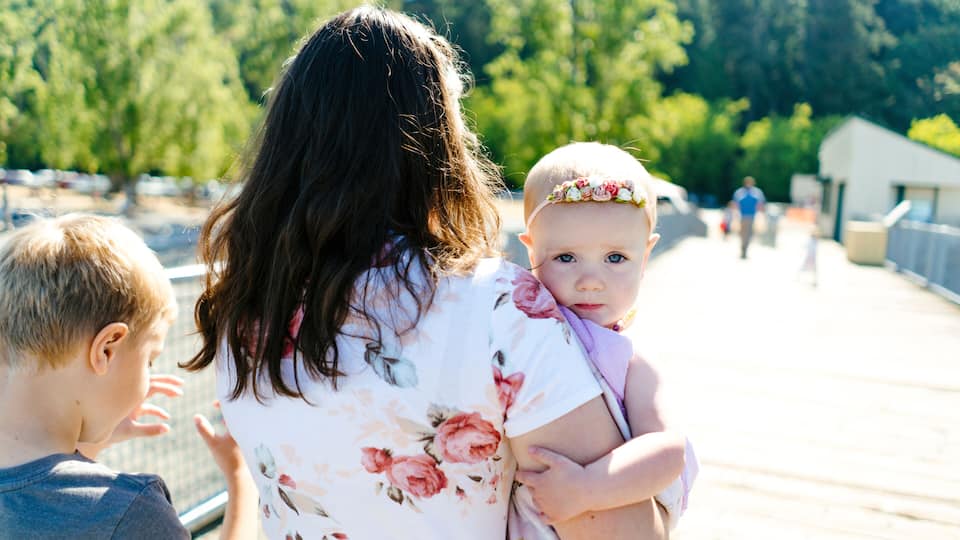 A woman holding a baby on a wooden boardwalk with trees and a building in the background.