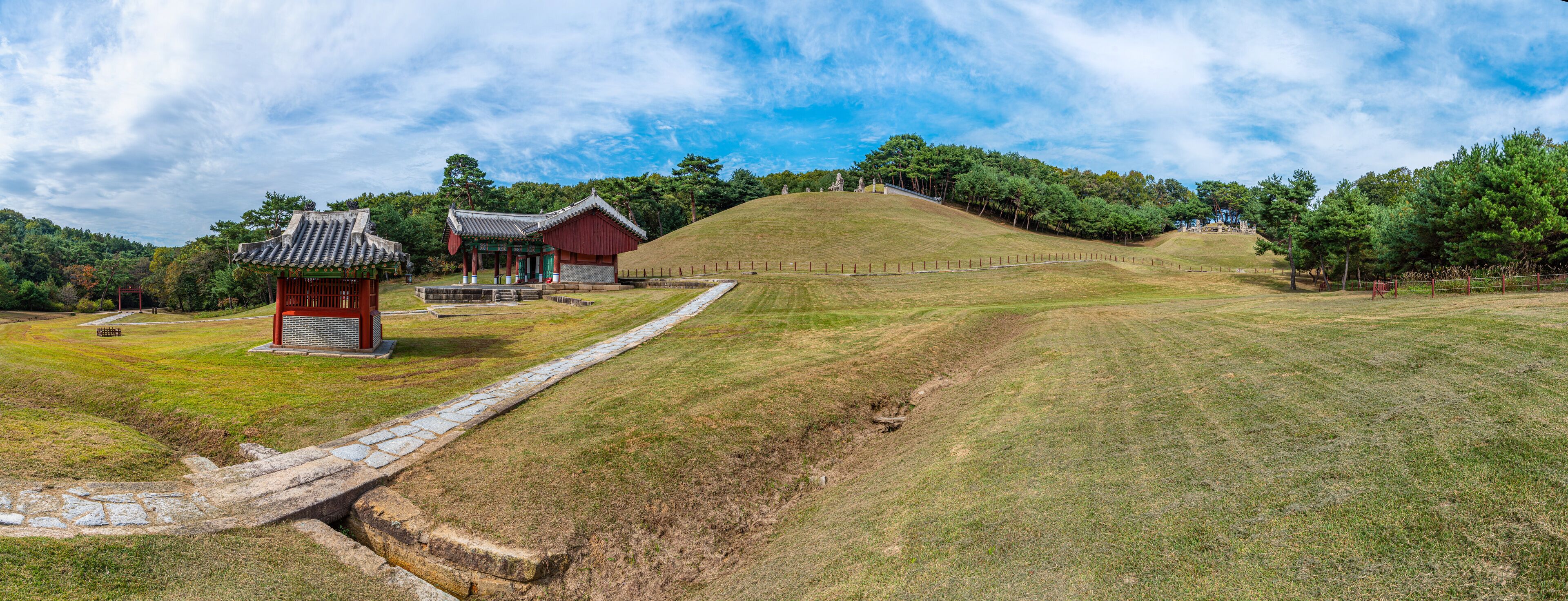 A traditional Korean pavilion, a red building, and a stone pathway in a park with a hill and trees.