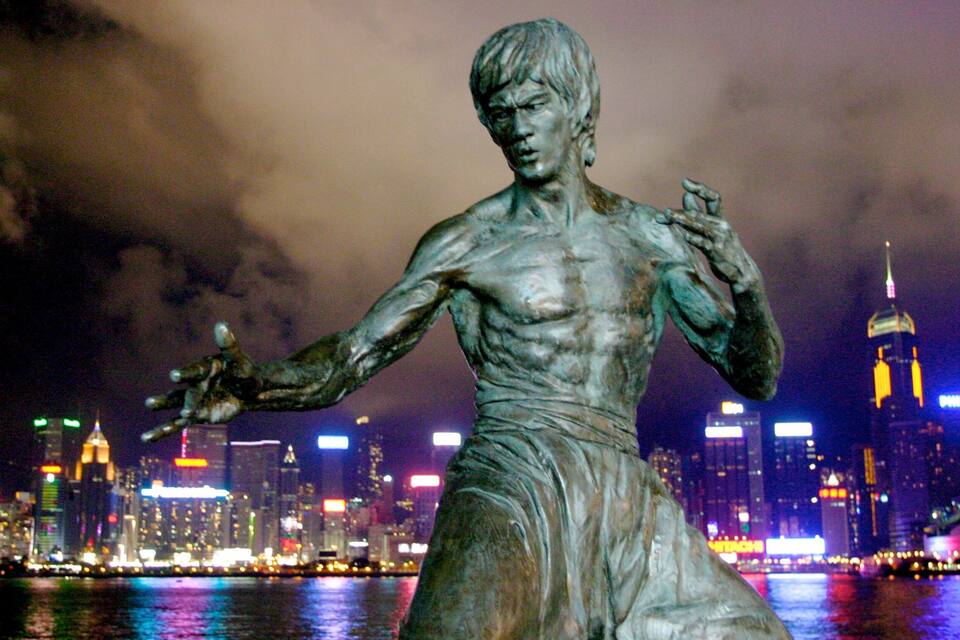 A statue of a muscular figure with outstretched arms, set against a city skyline with illuminated buildings and a body of water in the foreground.