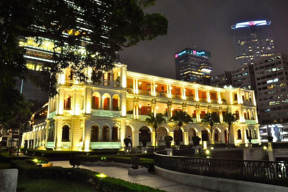 Um edifício histórico com janelas em arco e varanda, rodeado por um jardim bem iluminado e arranha-céus modernos ao fundo.