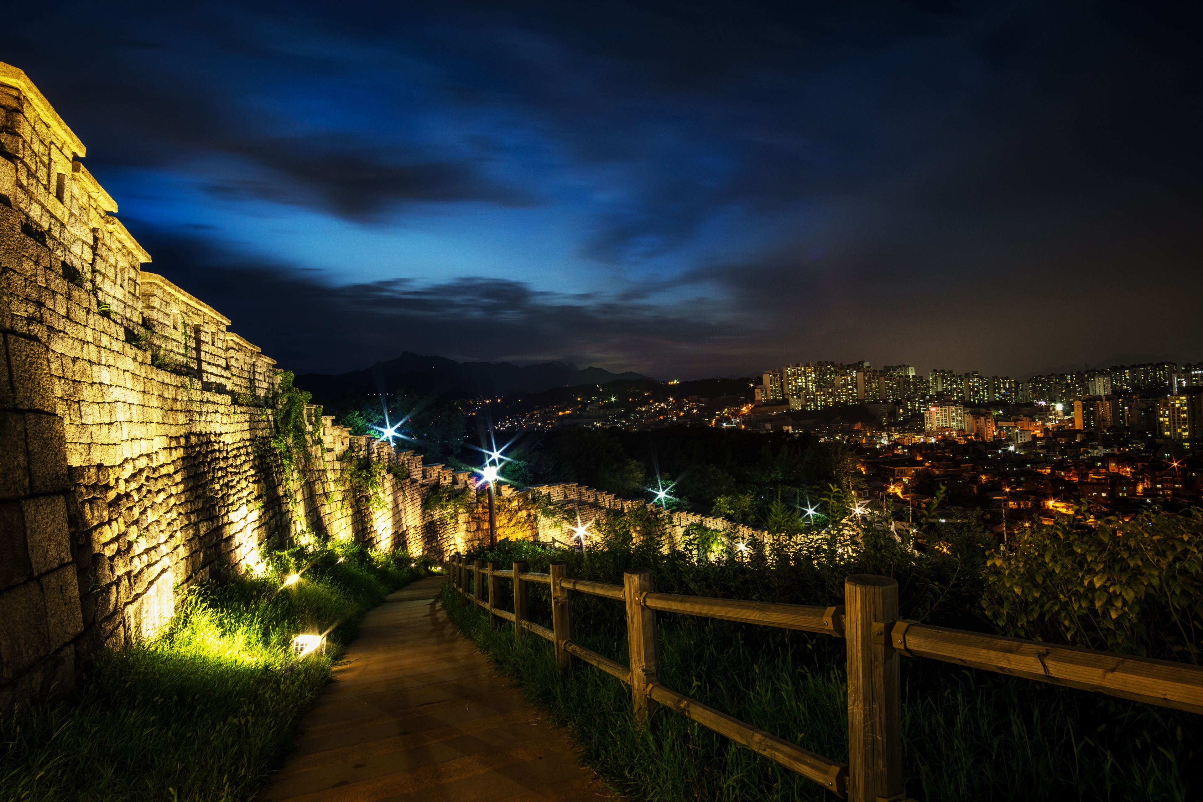 A nighttime cityscape with a historic stone wall and a pathway leading towards the city lights.