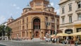 A historic building with ornate architecture, a prominent dome, and a street with pedestrians and cafes.