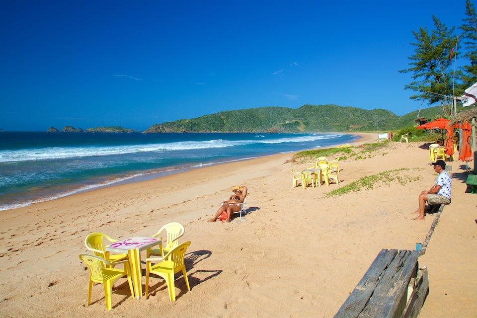 Uma praia com cadeiras e mesas amarelas, um banco de madeira e uma pessoa sentada na areia.
