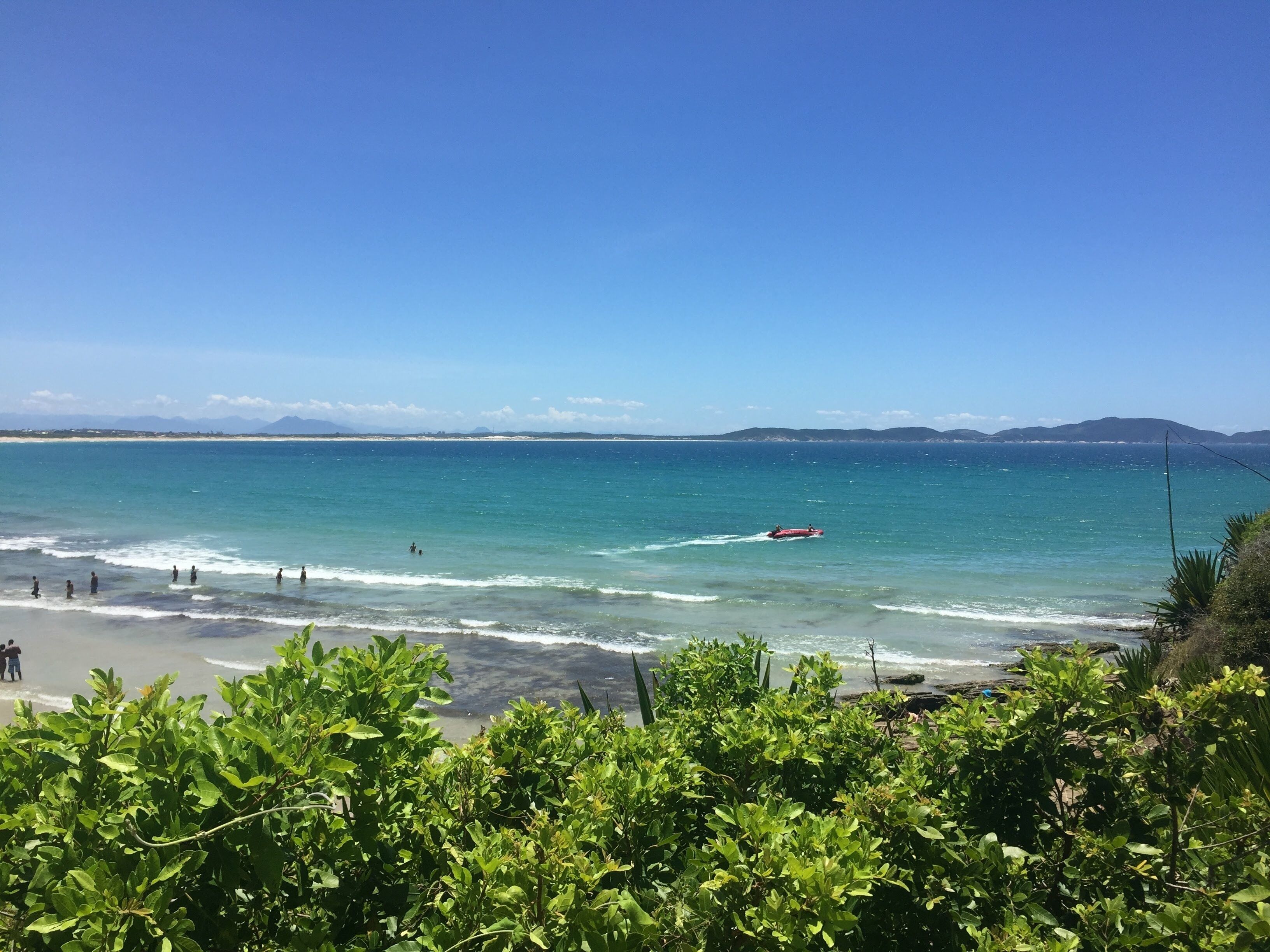 Uma praia com água azul cristalina, algumas pessoas na água e um barco vermelho ao longe.