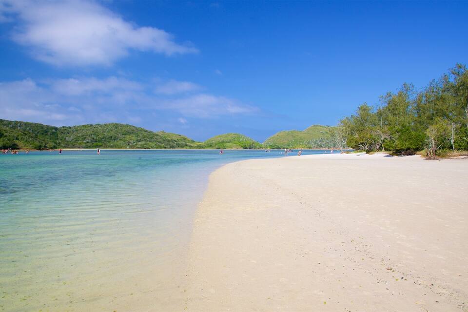 Uma praia com águas turquesa cristalinas, areal arenoso e colinas verdes ao fundo.