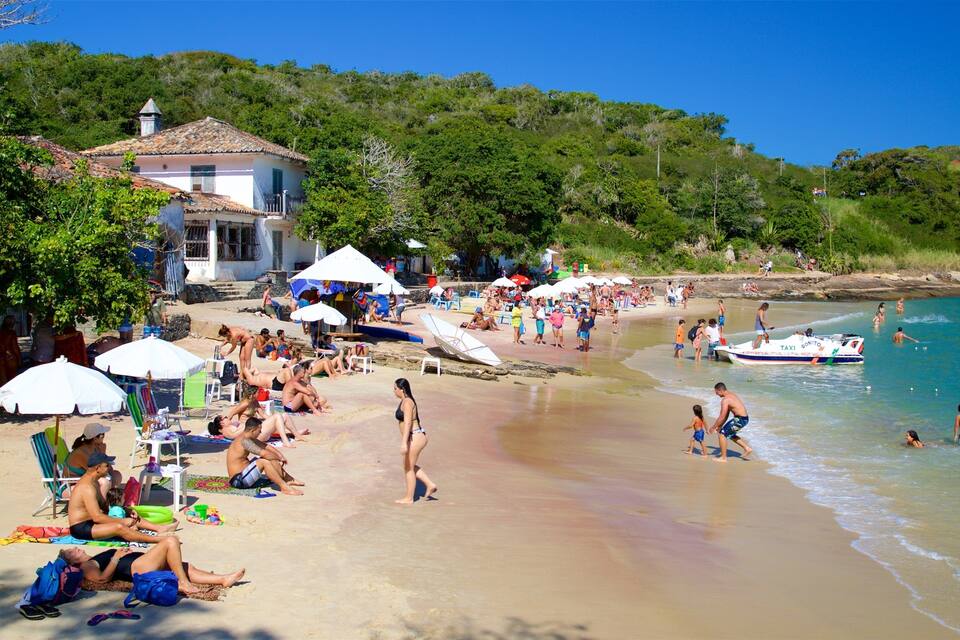 A crowded beach with people sunbathing, swimming, and playing. There are umbrellas, beach chairs, and a boat in the water.