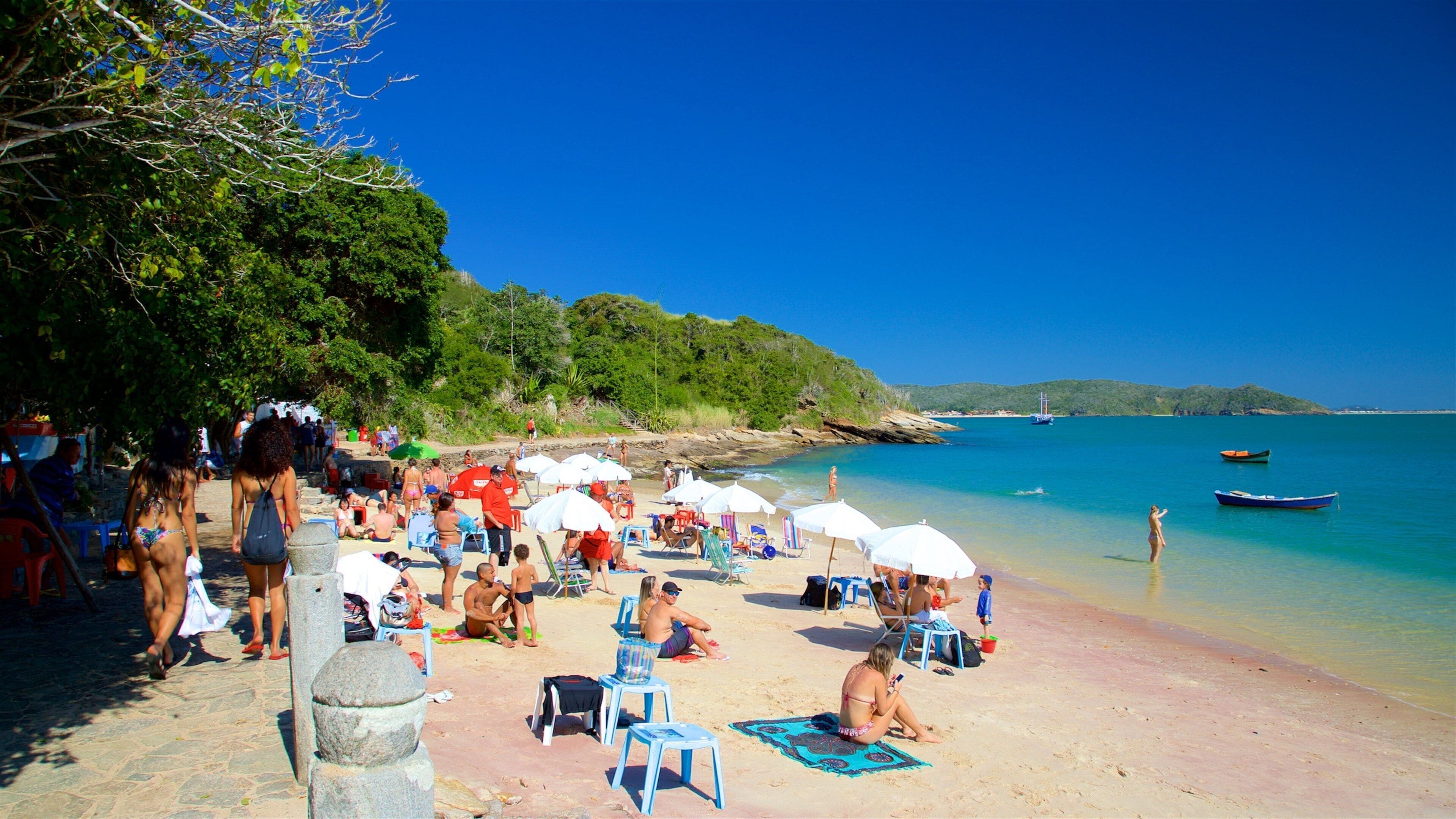 Una playa con gente tomando el sol, sombrillas y barcos en el agua.