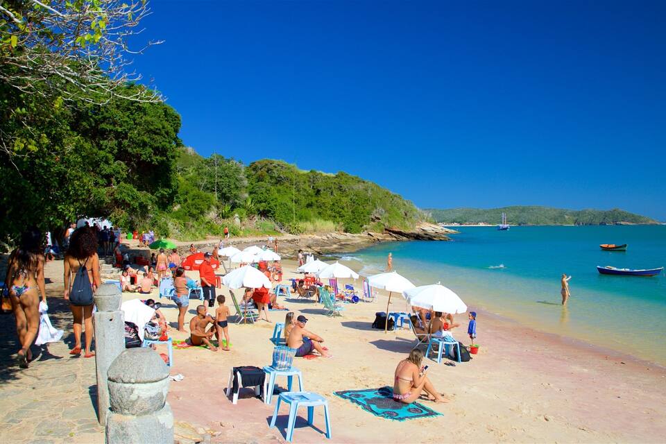 Una playa con gente tomando el sol, sombrillas y barcos en el agua.
