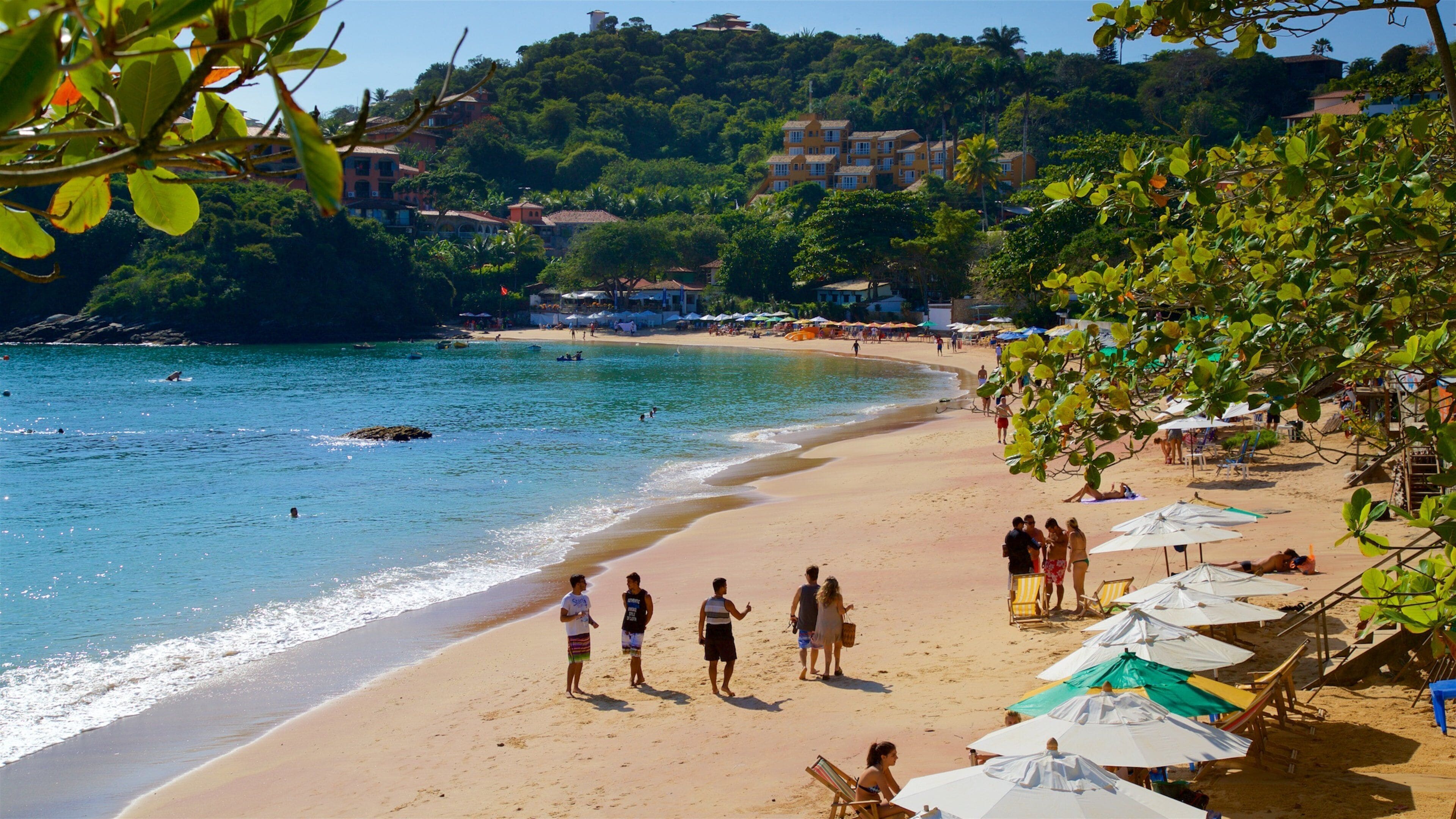 A beach with people walking along the shore, umbrellas, and buildings in the background.