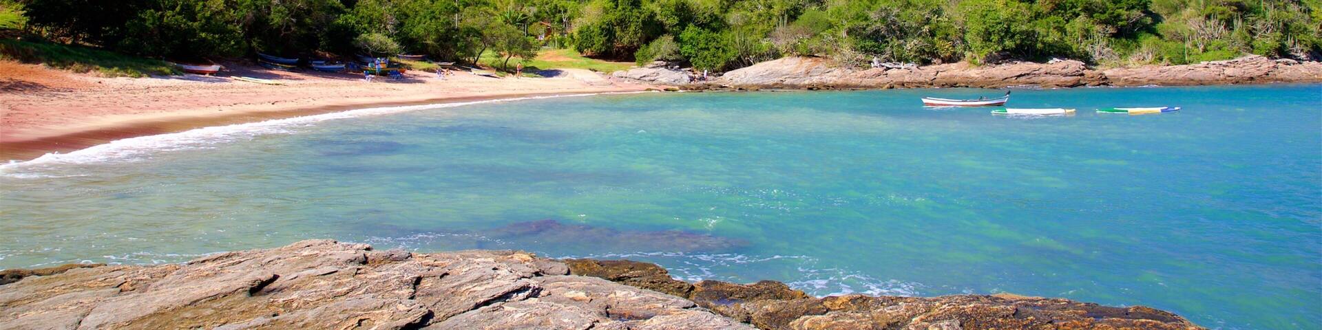 A clear turquoise beach with rocky foreground, lush green hills, and a few boats in the water.