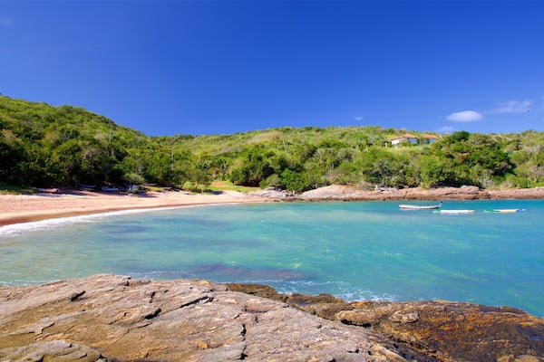 A clear turquoise beach with rocky foreground, lush green hills, and a few boats in the water.