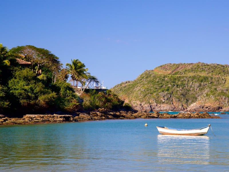 A calm coastal scene with a boat on the water, a rocky shoreline, and a hill with dense vegetation.